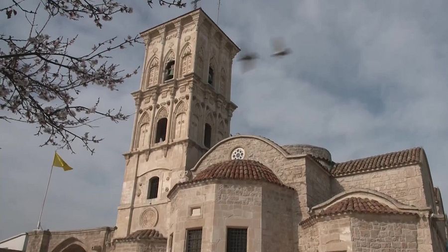 Iglesia bizantina de piedra con campanario cuadrado y techo rojo.  Bandera amarilla y árboles en flor visibles.  Destino turístico rural.