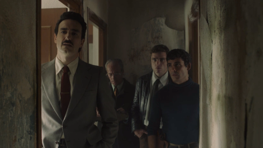 El comisario Velarde, interpretado por Miki Esparbé, con traje gris y corbata granate, observa una habitación en mal estado acompañado de tres hombres.