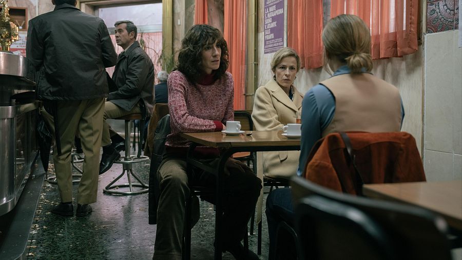 Tres mujeres charlan en una cafetería; dos hombres están en la barra.  Se observan detalles de sus ropas y dos tazas en la mesa.