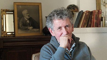 Retrato de Jean Michel Verne, con cabello canoso y rizado, sentado pensativo.  Al fondo, un retrato de Julio Verne y libros antiguos.