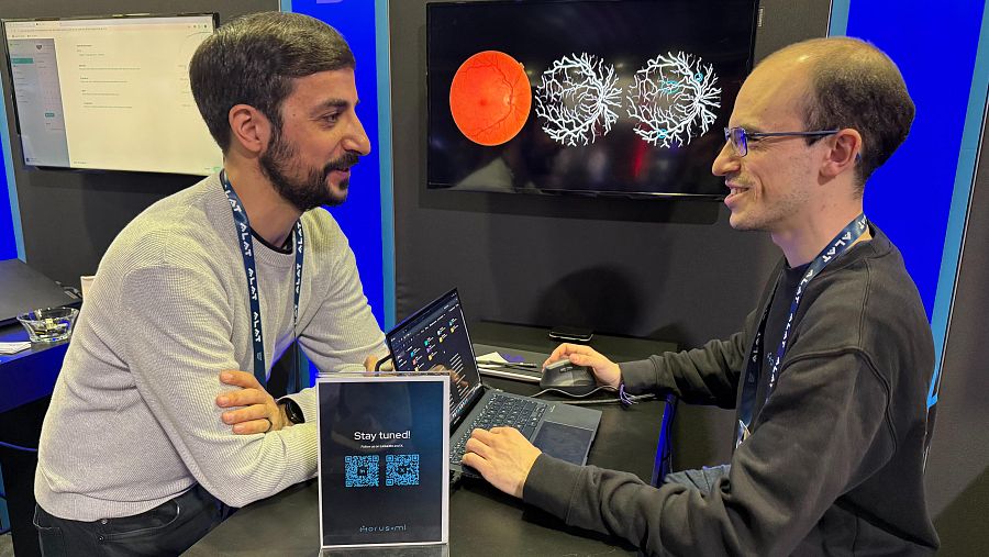 Presentación en un evento de un proyecto de detección precoz de una enfermedad, usando imágenes médicas y datos en un portátil. Dos hombres con acreditaciones explican el proyecto.