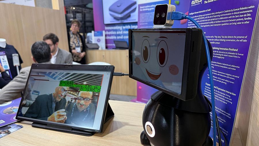 Robot social con pantalla expresiva, cámara y cable de conexión, sobre mesa de madera.  Ambiente de exposición.