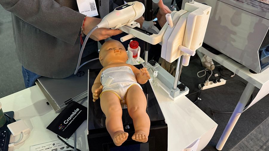 Dispositivo médico blanco con brazo articulado, usado sobre un muñeco de bebé por una persona.  Simula la detección no invasiva de infecciones mediante ultrasonidos e IA.