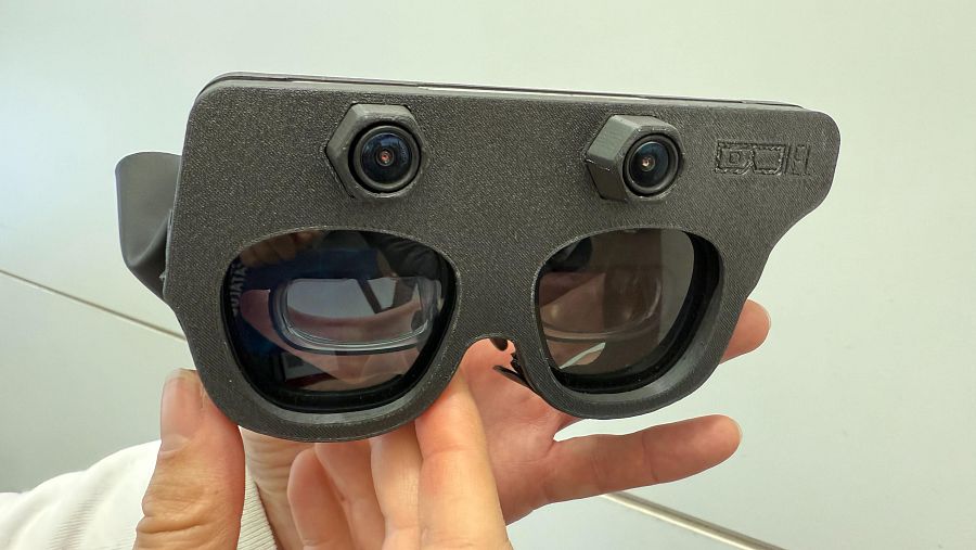 Manos sujetan unas gafas grises con dos lentes y cámaras, las Biel Glasses, un dispositivo tecnológico de asistencia visual.