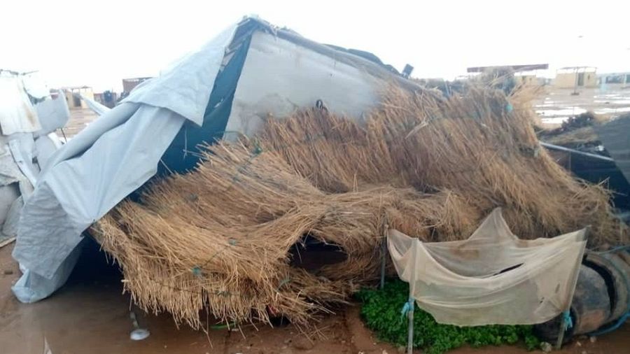 Precario refugio de refugiados, hecho de paja y lona rasgada, con mosquitera y charcos de agua. Se ven otras estructuras similares y un neumático.