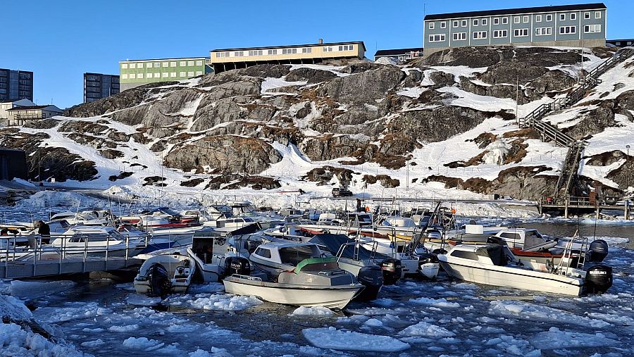 Puerto groenlandés con barcos entre bloques de hielo; edificios en una colina rocosa con escalera de madera.  El deshielo afecta la navegación.