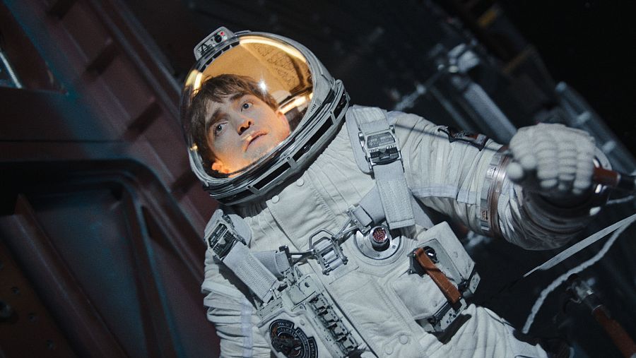 Astronauta en traje blanco con detalles naranja y gris, expresión preocupada, mirando hacia arriba en una nave espacial oscura y metálica.