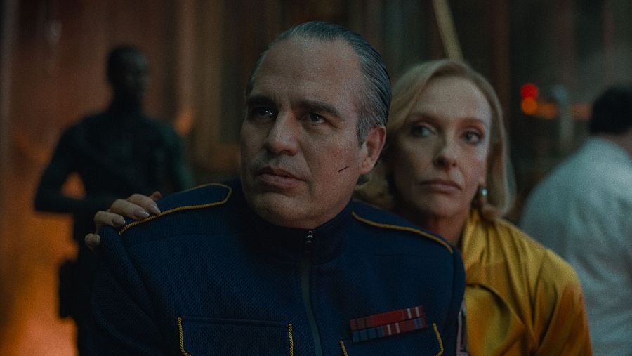 Oscar Isaac, con chaqueta azul y cicatriz, y Toni Collette, con ropa amarilla, comparten escena en un interior oscuro de 'Mickey 17'.  Expresiones serias.