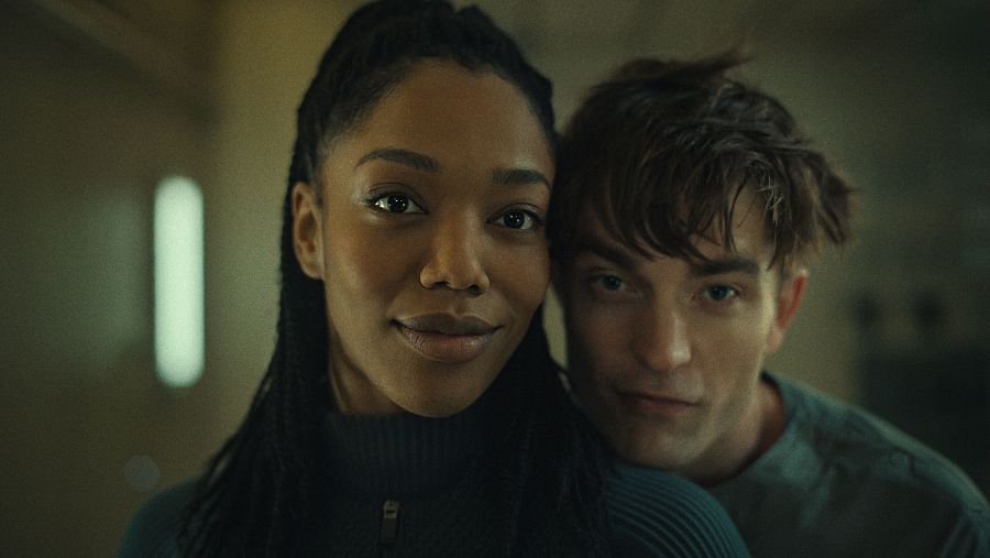 Fotograma de película: Primer plano de un hombre y una mujer, ella con trenzas y sonriendo, él con expresión seria. Iluminación tenue.