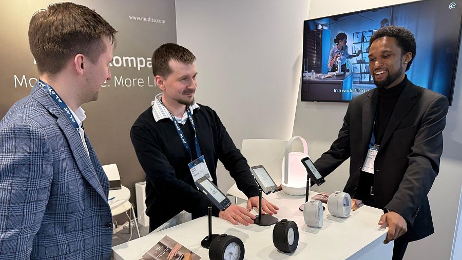 Tres hombres en un stand de exposición mostrando dispositivos electrónicos, con un cartel de la empresa y su sitio web visible.