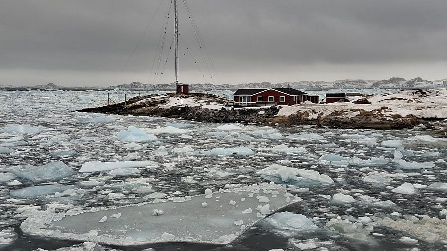 Casa roja con antena en costa groenlandesa, rodeada de hielo que evidencia el deshielo.  Cielo nublado y colinas nevadas al fondo.