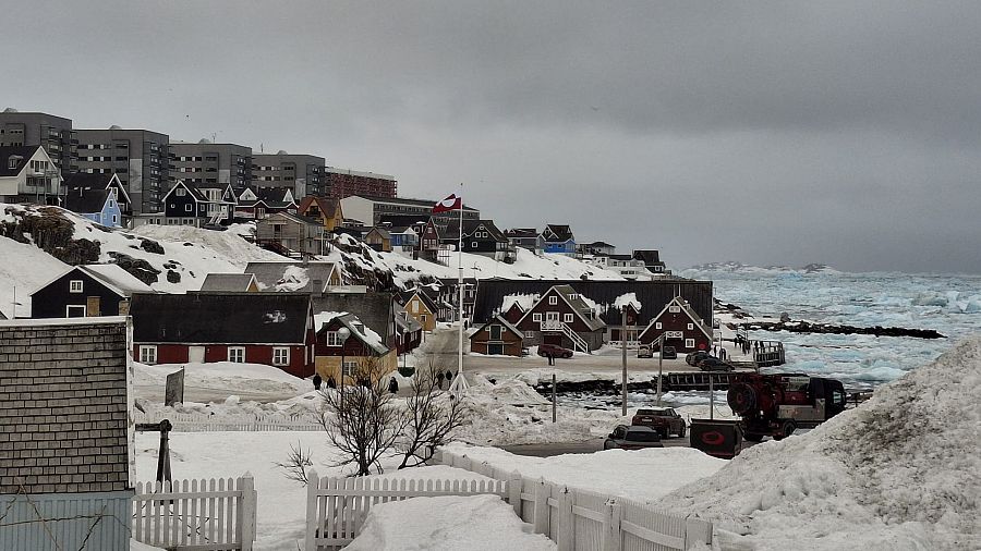 Ciudad costera nevada con casas de madera de colores vivos, edificios modernos y un quitanieves.  Mar parcialmente congelado al fondo.