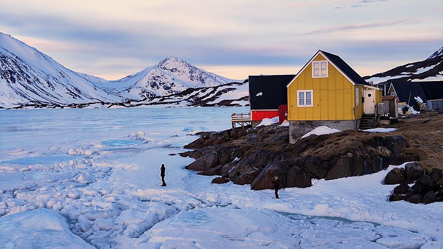 Lago congelado en paisaje invernal groenlandés, con casas y montañas nevadas. Dos personas con ropa oscura están cerca de la orilla helada.