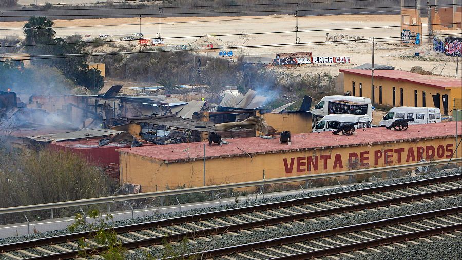 Daños por incendio o explosión en una empresa pirotécnica. Humo, estructuras dañadas y vehículos en techos. Se observa un letrero de 'VENTA DE PETARDOS'.