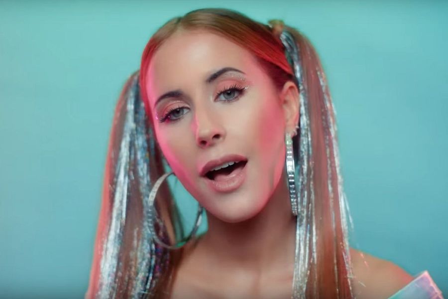 Retrato de la cantante Vicco con maquillaje brillante, coletas altas con reflejos plateados y pendientes grandes. Fondo turquesa.