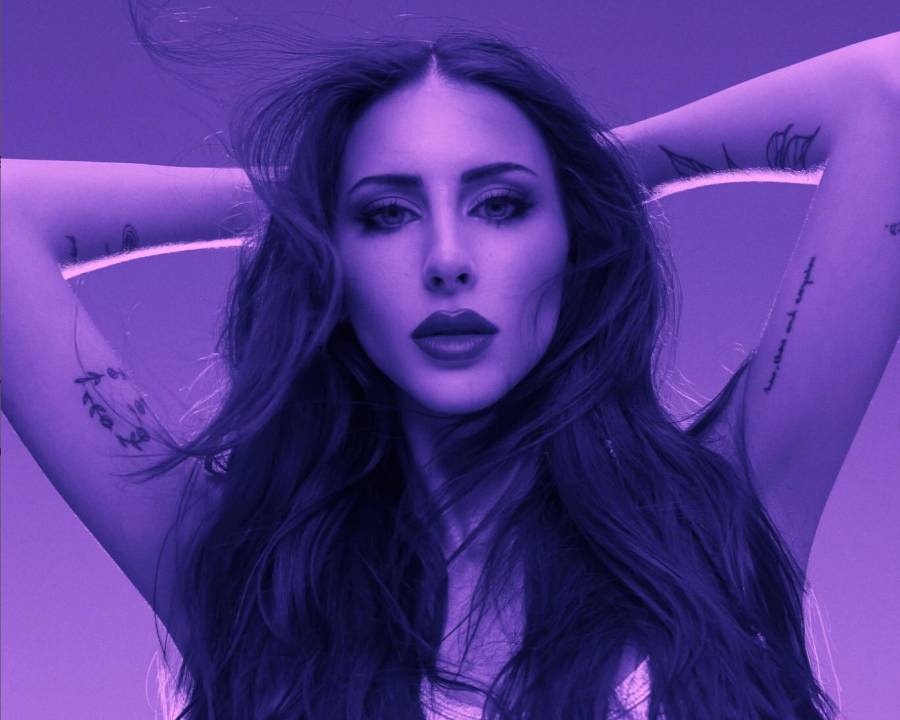 Cantante con cabello oscuro y tatuajes, en pose seria para foto promocional. Maquillaje visible y brazos flexionados sobre la cabeza.