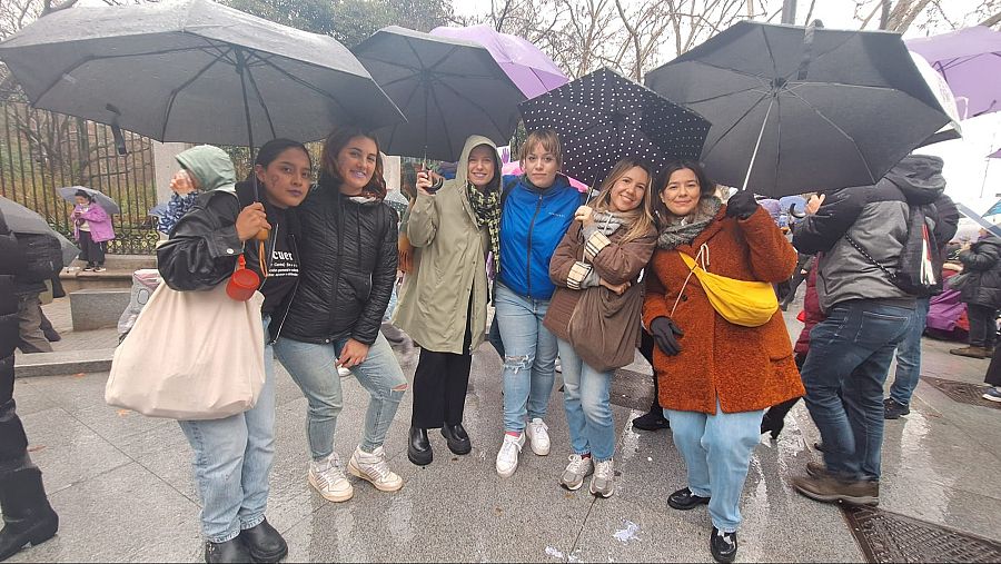 Grupo de mujeres con paraguas en día lluvioso. Una lleva un bebé. Ropa informal de otoño/invierno.  Fondo con más personas.