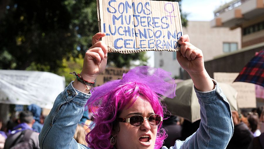 Manifestación 8M: Mujer con pancarta 'Somos mujeres luchando, no celebrando', peluca rosa y gafas de sol, expresión seria.