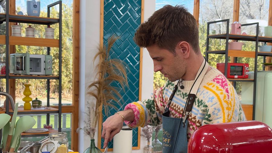 Actor en un programa de repostería, concentrado en una receta. Viste jersey navideño y delantal, en una cocina con azulejos verdes y microondas.