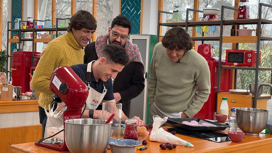 Exfutbolistas en un set de cocina de repostería, observando a otros concursantes.  Uno viste jersey mostaza, el otro verde.