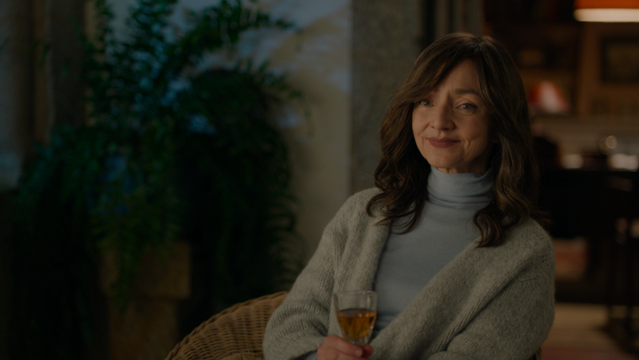 Actriz con jersey azul y chaqueta gris, sosteniendo una copa, en escena teatral.  Expresión serena, fondo desenfocado.