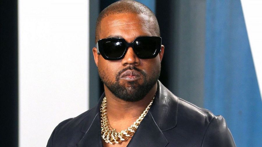 Imagen de Kanye West con cabeza rapada, gafas oscuras, cadena de oro y chaqueta oscura; expresión seria.