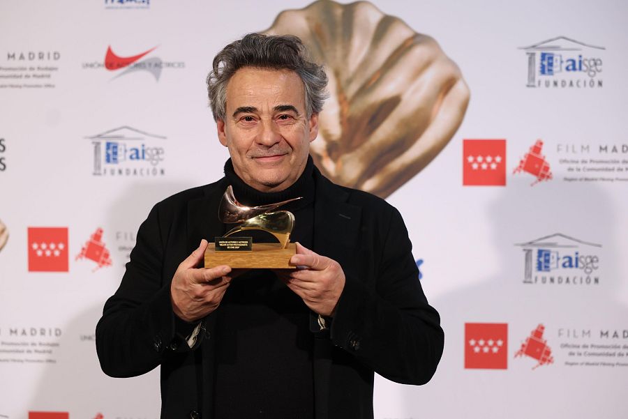 Eduard Fernández, con chaqueta y jersey negros, sostiene un premio dorado en un photocall con logotipos de Madrid y otras instituciones.  Muestra una expresión de satisfacción.