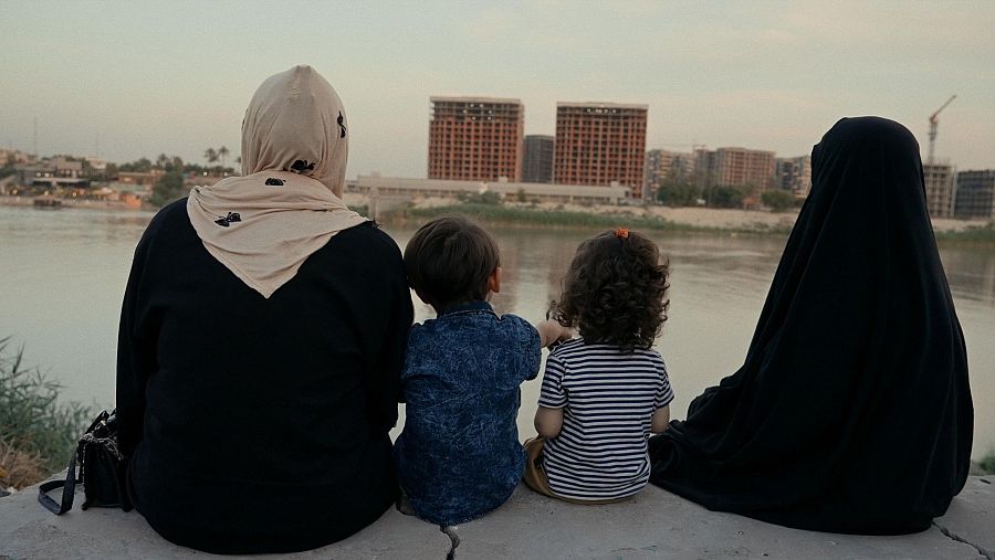 Mujer con hiyab y dos niños a la orilla de un río; al fondo, construcciones.  La imagen refleja la precariedad económica de la familia.