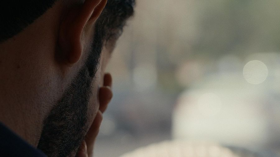 Hombre con barba, de perfil, mano en mejilla, mirando a través de un cristal difuminado.  Postura pensativa que refleja una situación difícil.