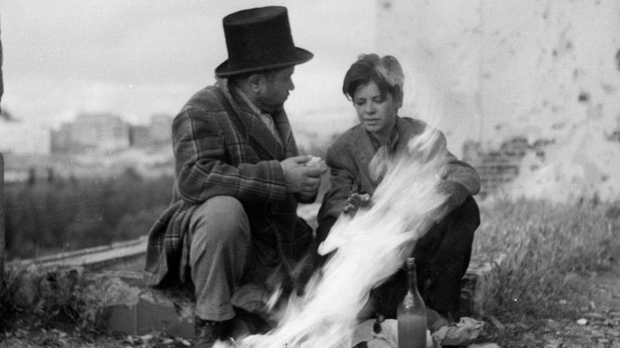 Adulto con sombrero de copa y niño junto a fogata en escena en blanco y negro.  Entorno desolado con edificios al fondo.  Sensación de pobreza.