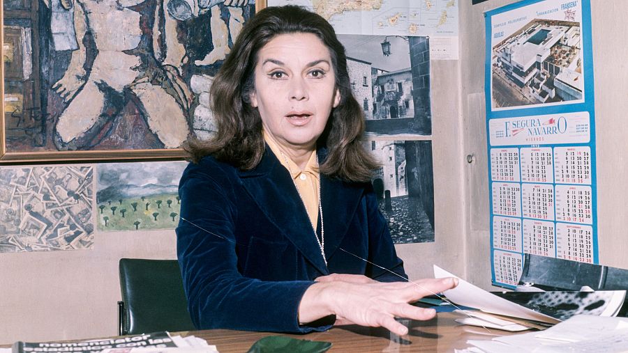 Directora de cine sentada en un escritorio, con chaqueta azul y camisa amarilla, gesticulando.  En la pared, cuadros, fotos y un calendario. Sobre el escritorio, papeles y una revista.