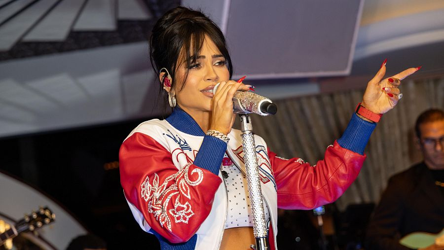 Cantante con chaqueta bomber roja y blanca, micrófono en mano, actuando en un escenario con músicos de fondo.  Pelo recogido, accesorios visibles.