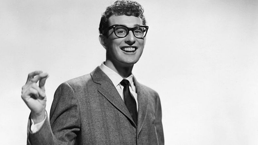 Retrato en blanco y negro de Buddy Holly; sonriente, con gafas y traje, gesticula mientras mira a la cámara.