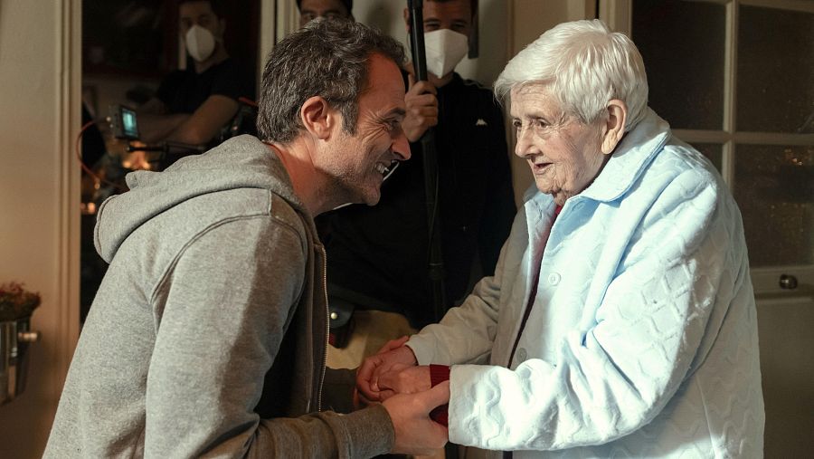 Hombre y anciana sonríen, tomados de la mano, en una escena de rodaje.  El equipo de filmación, con mascarillas, trabaja en segundo plano.