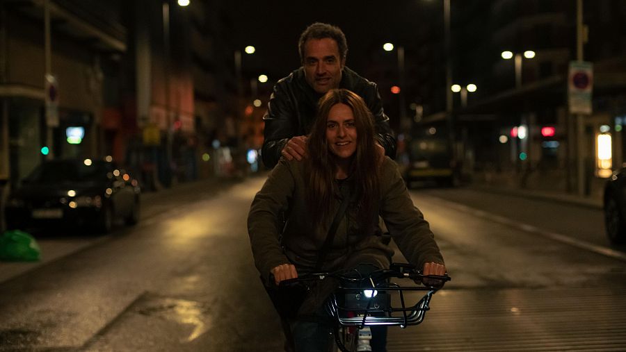 Itziar Ituño y Daniel Guzmán, en escena nocturna de película, ella en bicicleta, él apoyándola. Sonríen a cámara.