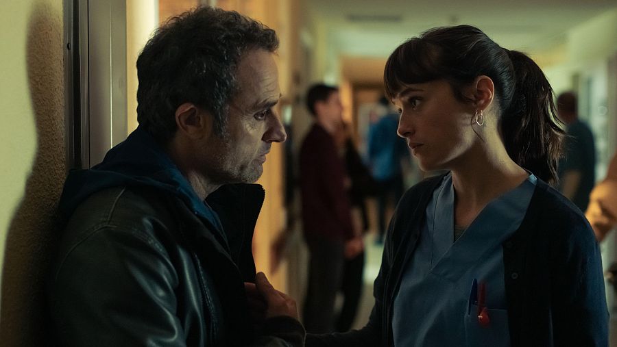 Hombre con chaqueta de cuero y mujer con uniforme sanitario mantienen una conversación tensa en un pasillo estrecho y con iluminación tenue.