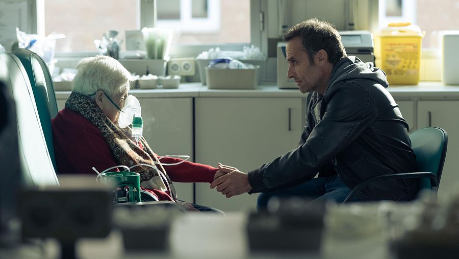 Escena conmovedora en un hospital: Daniel Guzmán sostiene la mano de Rosario García, quien lleva una máscara de oxígeno.  Ambiente íntimo y tenso.