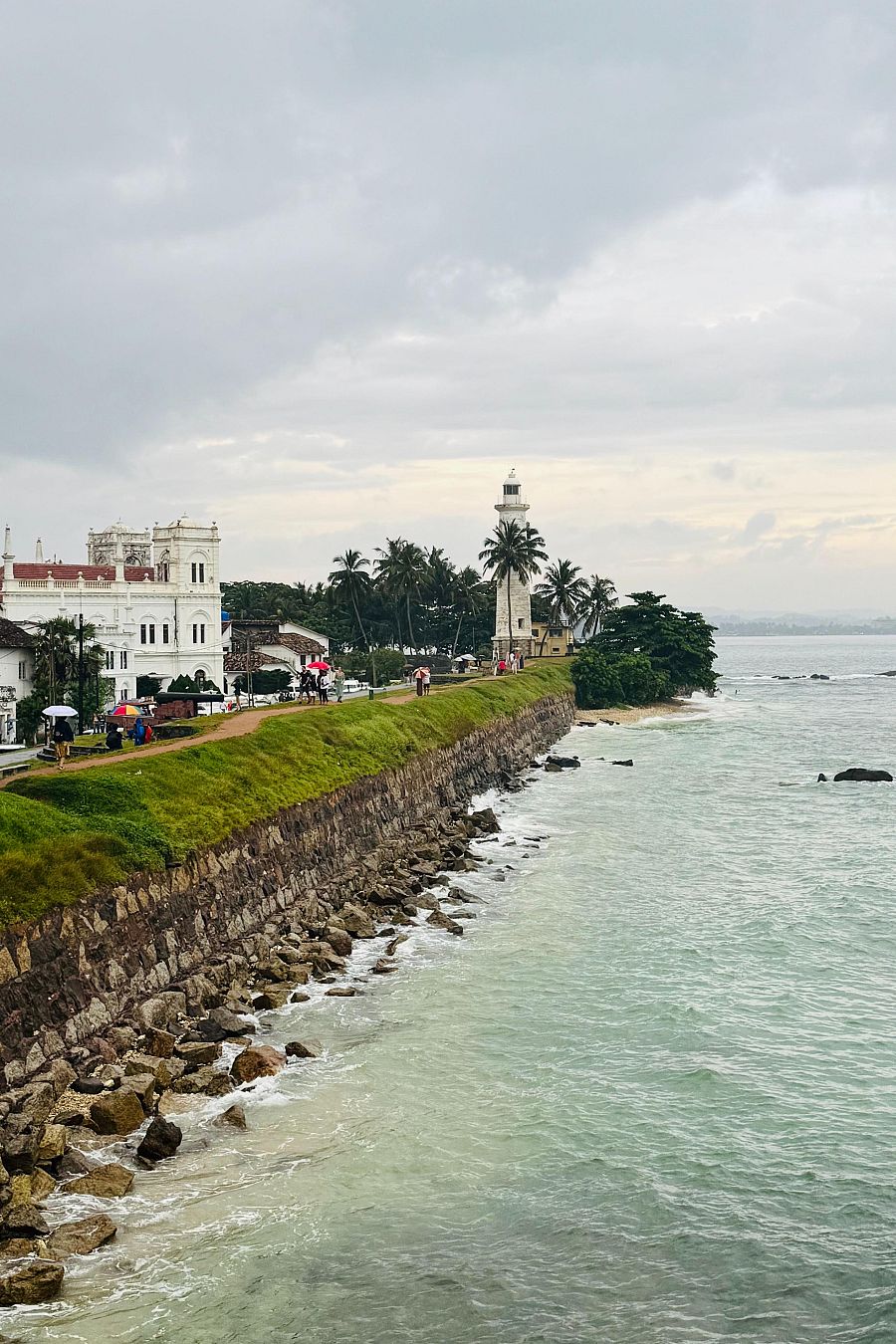 Faro blanco en costa, edificios coloniales con palmeras, personas paseando bajo cielo nublado.  Mar con olas suaves.