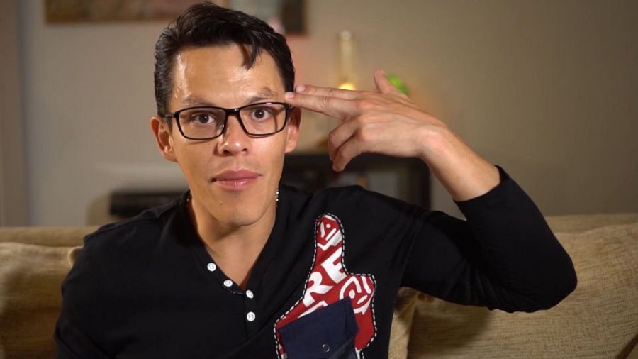 Joven con gafas y camiseta negra con bolsillo azul marino, sentado en un sofá beige, simula una pistola con la mano derecha apuntando a su sien; expresión seria.