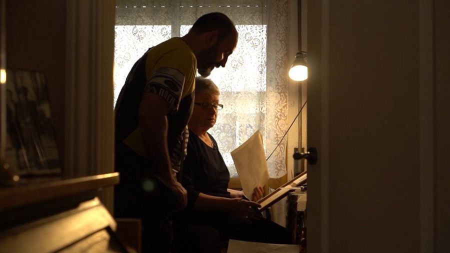 Un hombre y una mujer, posiblemente madre e hijo, revisan un documento.  Ella señala un objeto rectangular mientras él observa. Ambiente tranquilo y doméstico.