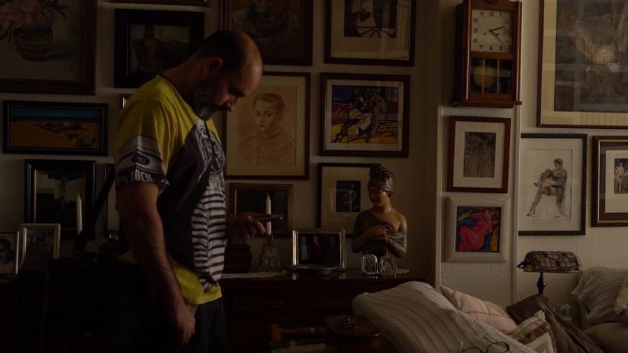 Hombre de mediana edad con cabeza rapada y barba, mirando su móvil en una habitación con cuadros, reloj y lámpara. Fotograma de documental.