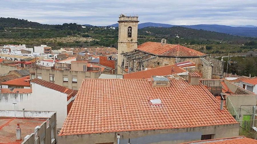 Vista panorámica de Càlig desde una torre, con tejados de tejas naranjas, una iglesia y un paisaje montañoso al fondo.