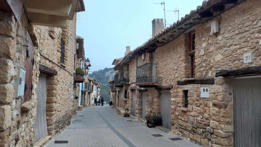 Calle estrecha con edificios antiguos de piedra, algunos con balcones y antenas en los tejados. Se ven números en las fachadas y farolas.
