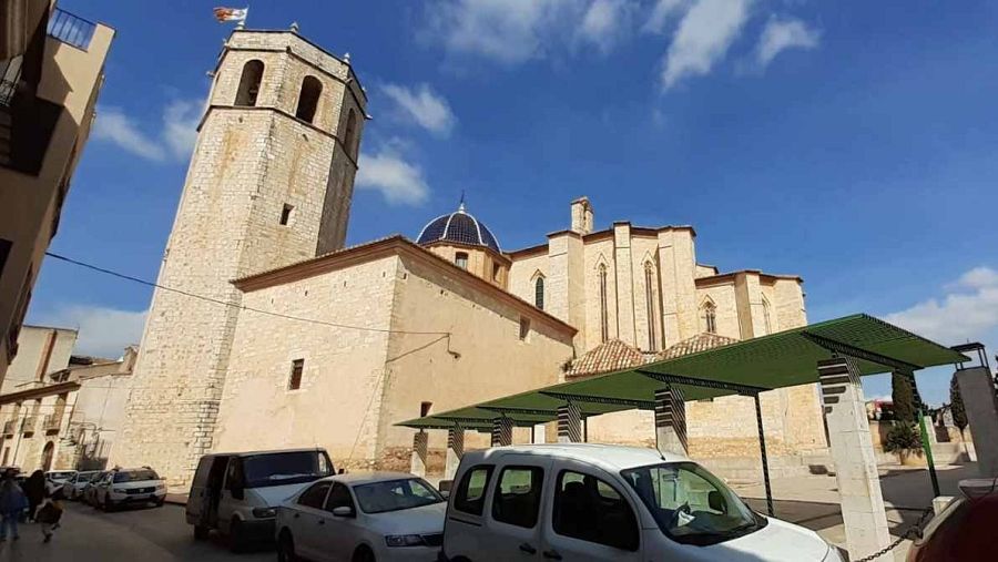 Iglesia de Sant Mateu, gótico valenciano, con torre octogonal, cúpula y detalles arquitectónicos góticos.  Aparcamiento moderno y coches en la calle adyacente.