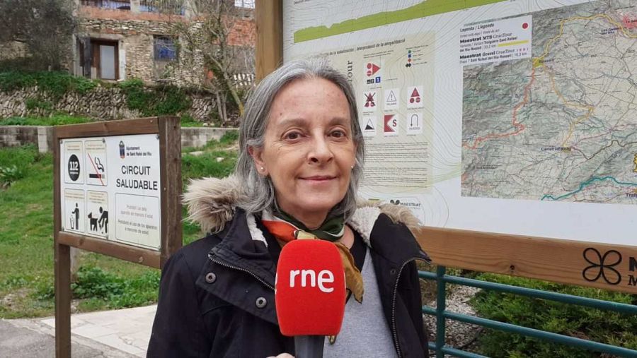 Técnica de turismo informa sobre el Gran Tour del Maestrat, frente a paneles con mapa de rutas y pictogramas de circuito saludable.  Micrófono RNE.