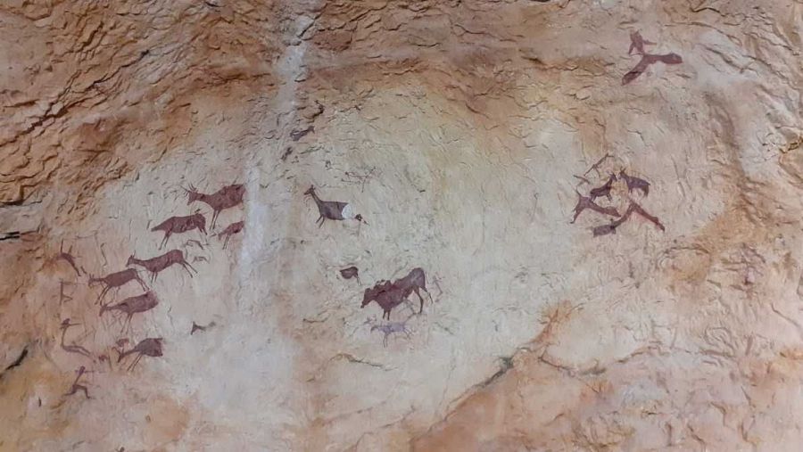 Pinturas rupestres rojizas en pared rocosa muestran figuras humanas y animales (ciervos, posiblemente caballos o bóvidos) en una escena que parece representar una cacería.