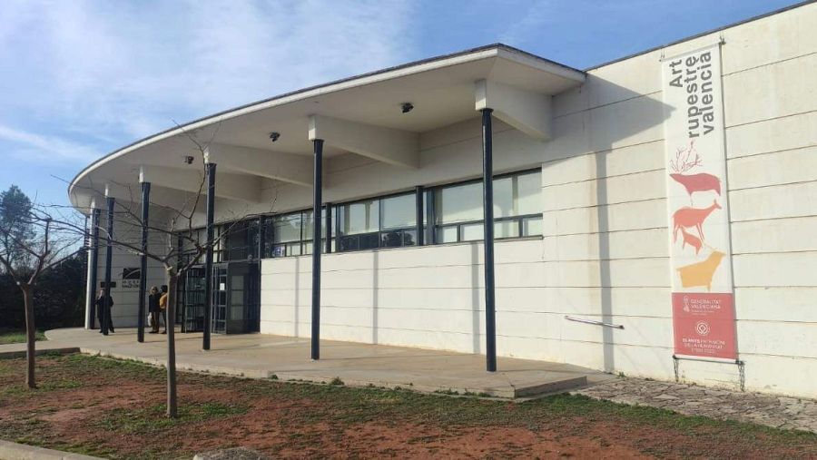 Moderna fachada de museo con entrada curva y cartel anunciando 'Art rupestre valencia' con figuras de animales prehistóricos.  Entorno con tierra rojiza y vegetación.