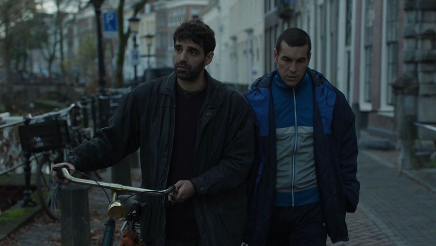 Dos hombres caminando por una calle junto a un canal en una ciudad europea, uno con una bicicleta. Árboles sin hojas, atmósfera sombría.
