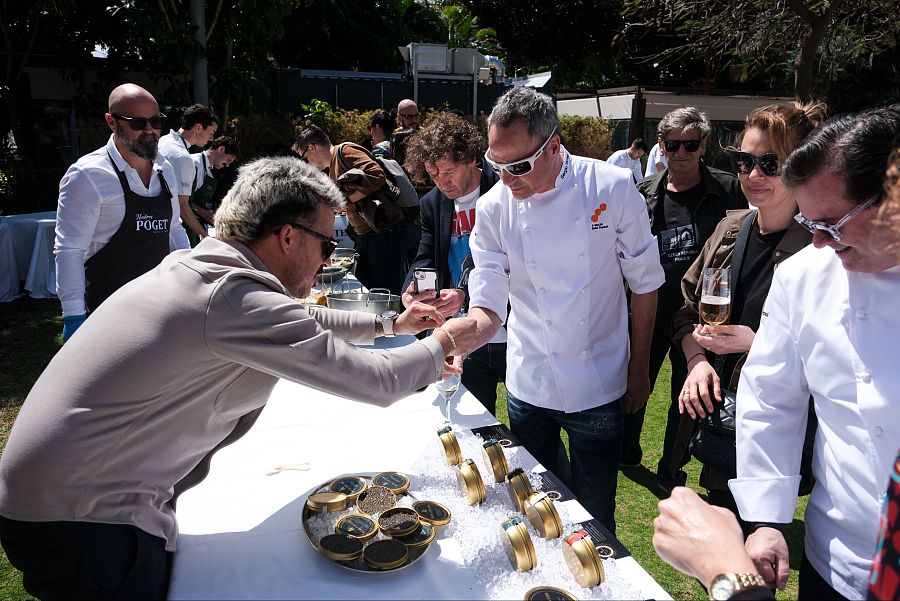 Degustación de caviar en evento al aire libre. Personas, entre ellas cocineros, prueban pequeñas latas doradas sobre hielo en una mesa blanca. Ambiente soleado.