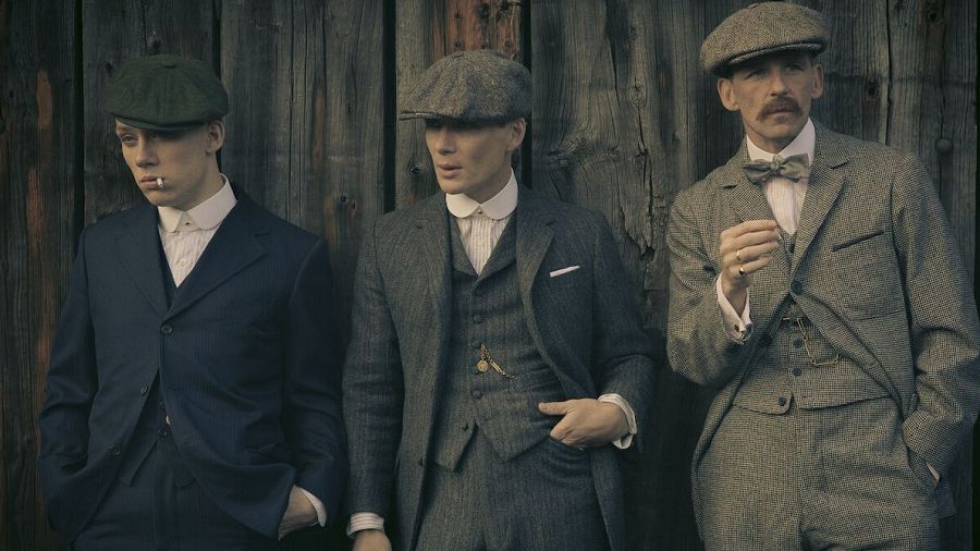 Tres hombres en trajes de época, uno hablando, los otros escuchando, apoyados en una pared de madera.